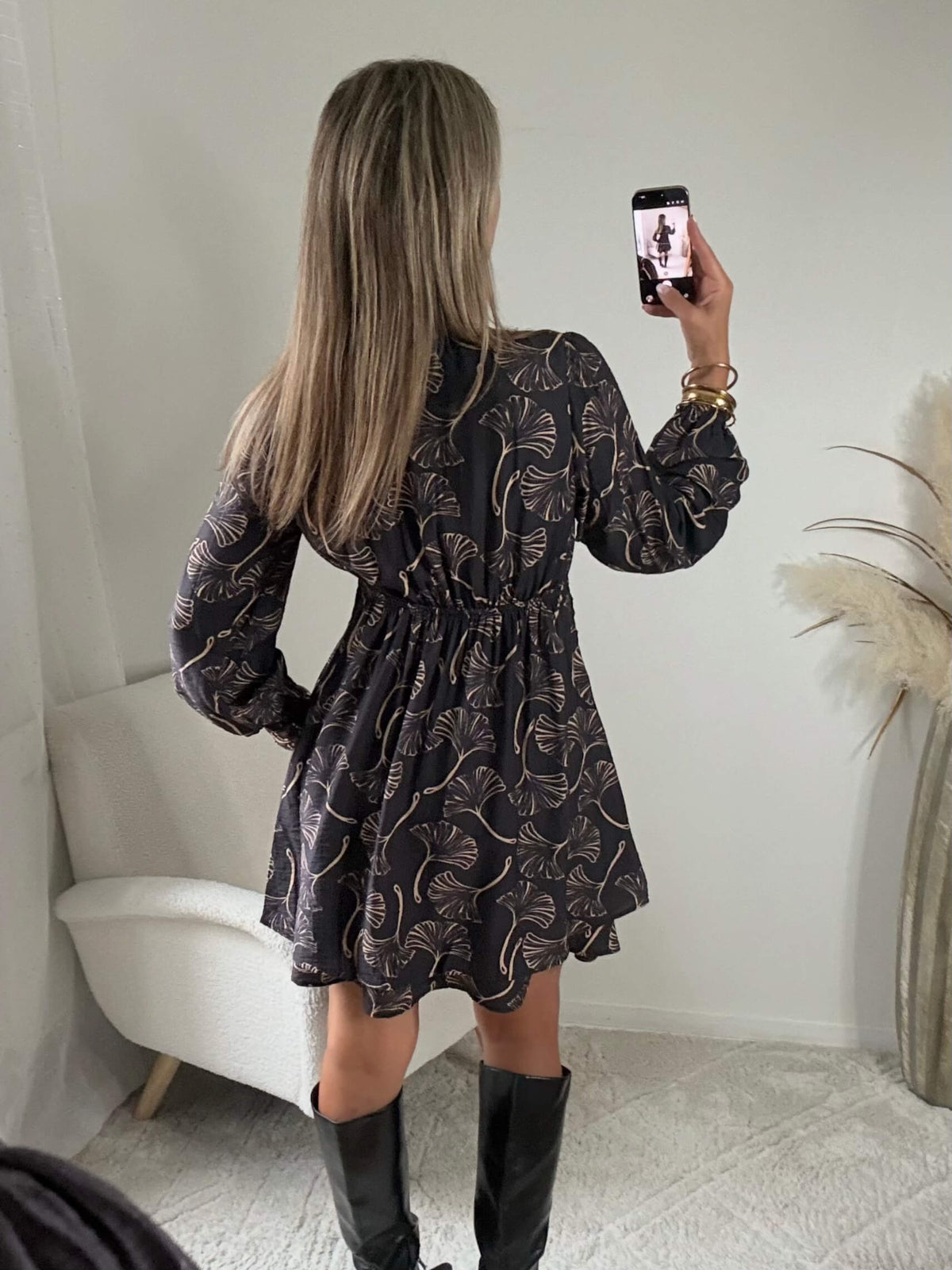 Robe courte noire Zéline du S au 4XL à motifs de feuillage dorés, décolleté croisé en V et manches longues, portée avec des bottes noires.