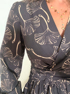 Robe courte noire Zéline du S au 4XL à motifs de feuillage dorés, décolleté croisé en V et manches longues, portée avec des bottes noires.