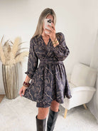 Robe courte noire Zéline du S au 4XL à motifs de feuillage dorés, décolleté croisé en V et manches longues, portée avec des bottes noires.